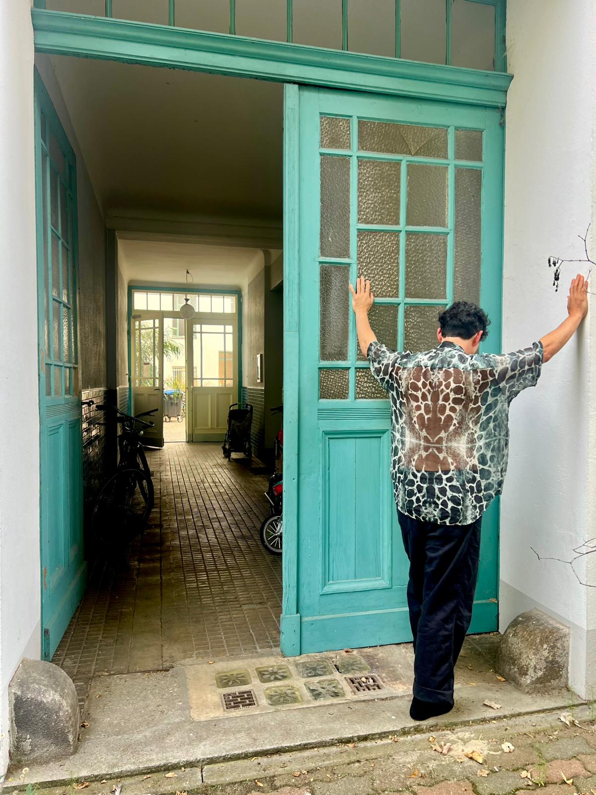 Model standing in front of a teal door with arms outstretched, while wearing Serengeti Stone shirt by Leon Dream.